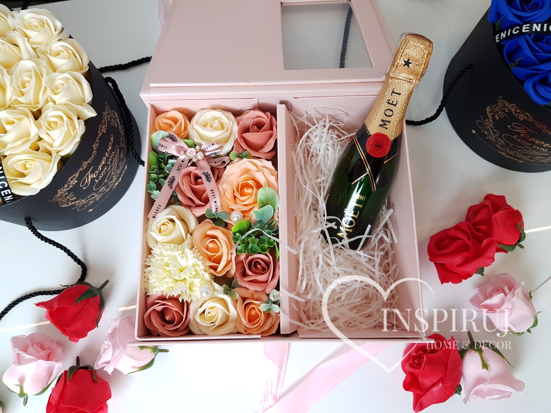 Elegancki kosz prezentowy z mydlanych róż w odcieniach kremu, brzoskwini i różu, uzupełniony o małą butelkę Moët & Chandon, umieszczony w różowym pudełku z okienkiem, otoczony luźnymi, czerwonymi...