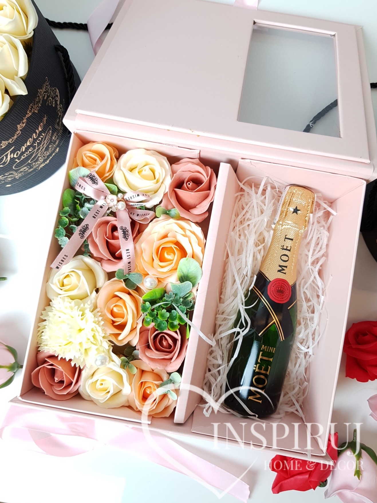 Otwarty różowy box prezentowy z mydlanymi różami w odcieniach brzoskwini i bieli, przewiązany wstążką z napisem 'Best Wishes', obok mini butelka szampana Moët w białym wypełniaczu.