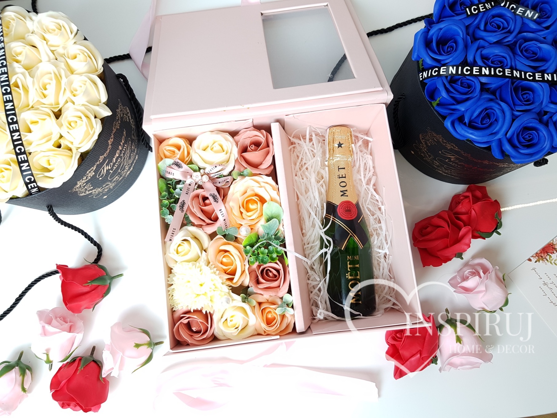 Otwarty, jasnoróżowy box prezentowy z okienkiem, wypełniony sztucznymi różami w odcieniach brzoskwini i beżu, oraz miniaturową butelką szampana Moët, otoczony koszami z białymi i niebieskimi różami...
