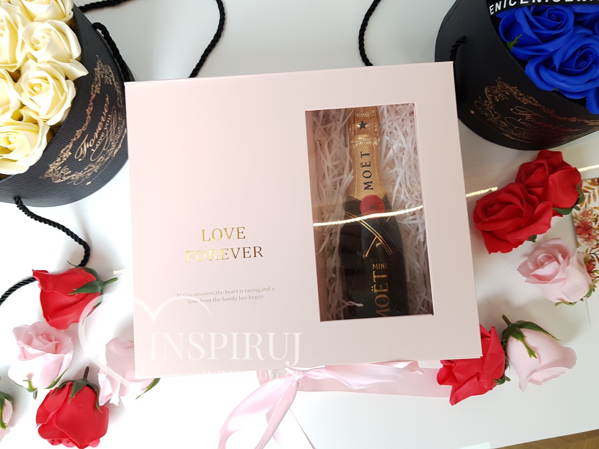 Różowy pudełko z napisem 'LOVE FOREVER' i okienkiem prezentującym mini butelkę szampana Moët, otoczone kompozycjami z mydlanych róż w odcieniach czerwieni, różu, bieli i błękitu, umieszczonych...