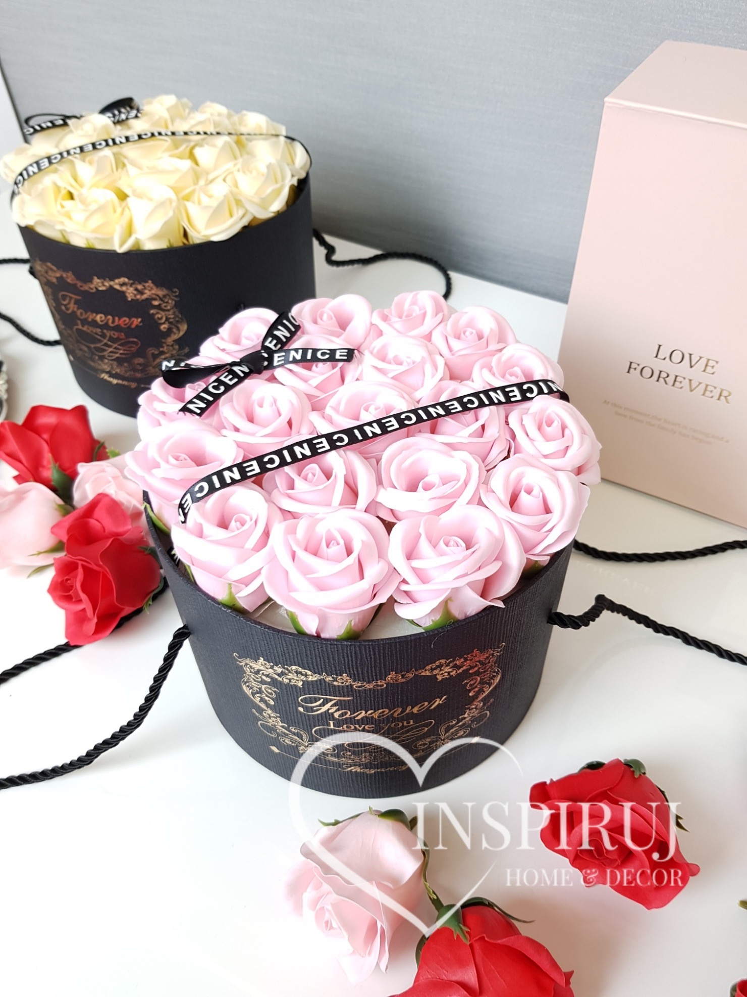Różowe i kremowe róże z mydła w czarnych, okrągłych pudełkach z napisem 'Forever' oraz wstążką z napisem 'NICENICENICE'. Pojedyncze, czerwone i różowe róże leżą obok.