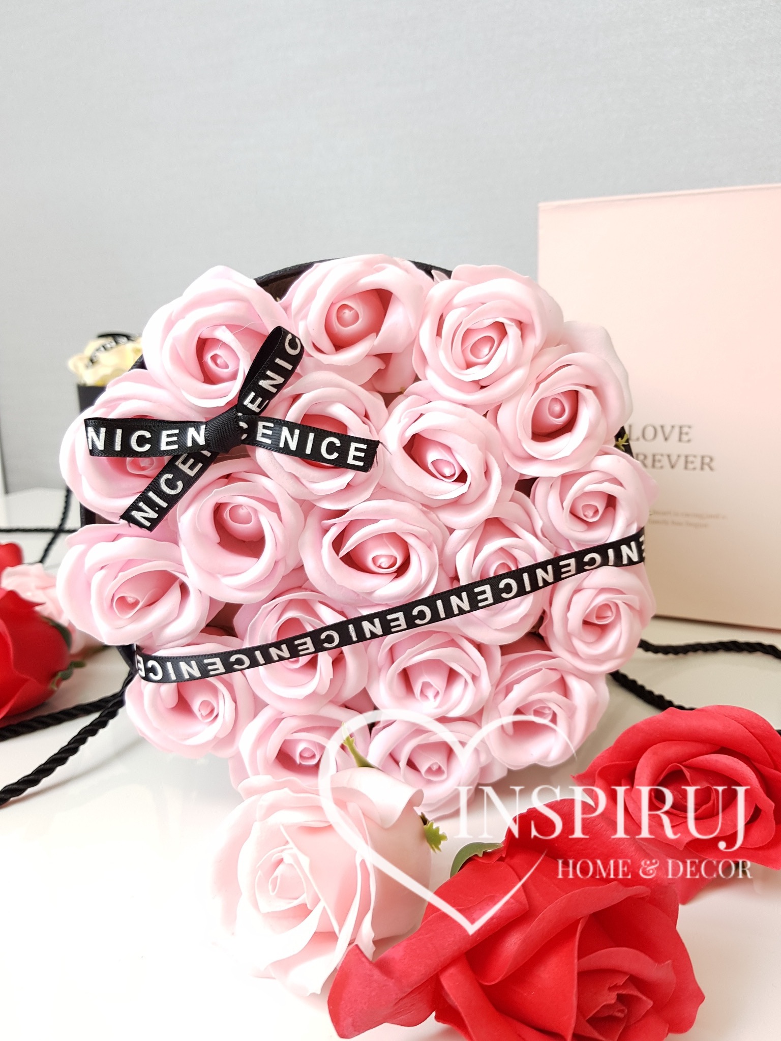 Różowy kosz róż z czarną wstążką 'NICE NICE', czerwone róże dekoracyjne obok, na białym tle.