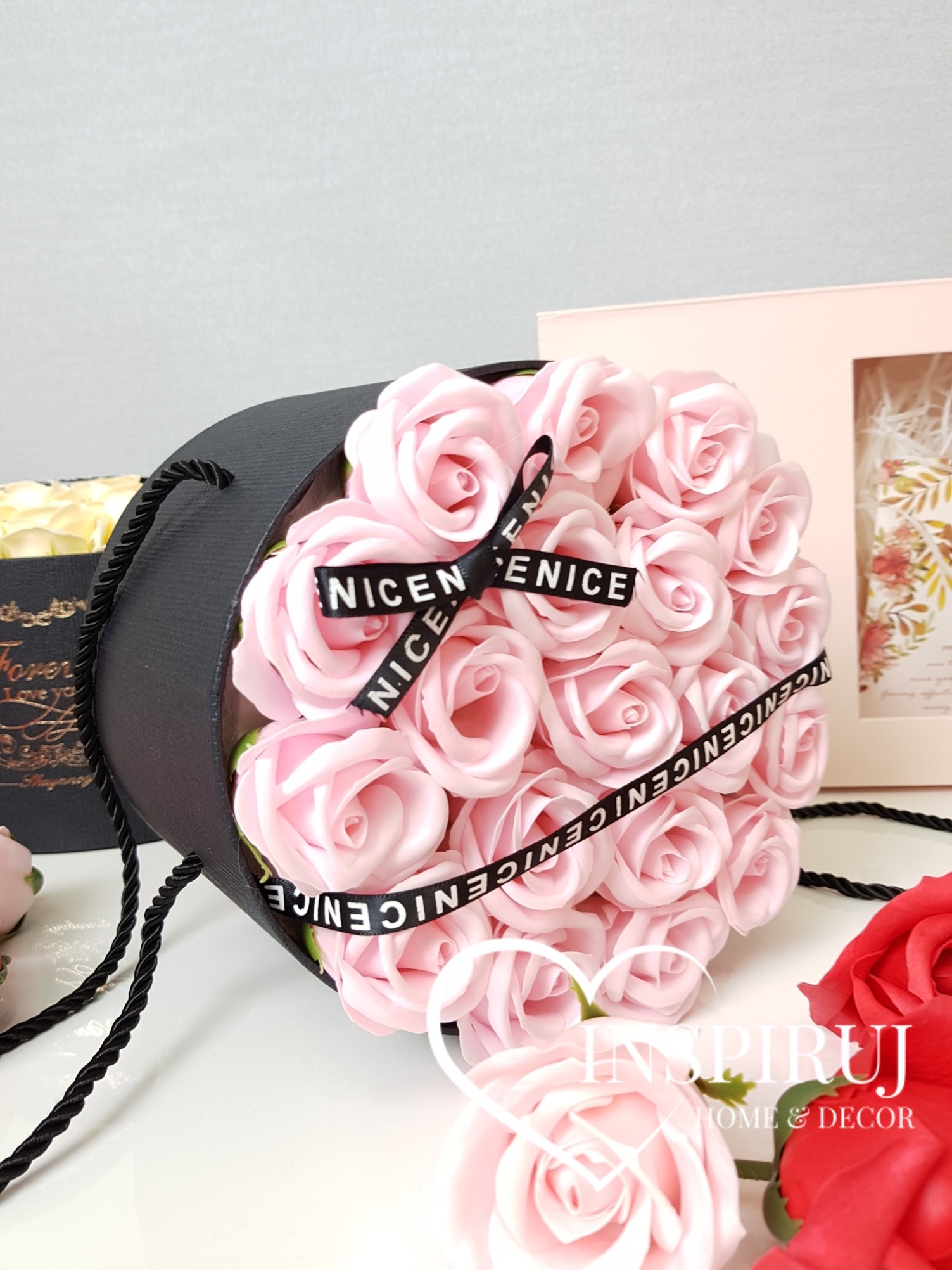Okrągłe, czarne pudełko z jasnoróżowymi różami z materiału, przewiązane czarną wstążką z napisem 'NICE', obok dekoracyjne pudełko z suszem kwiatowym i czerwona róża.