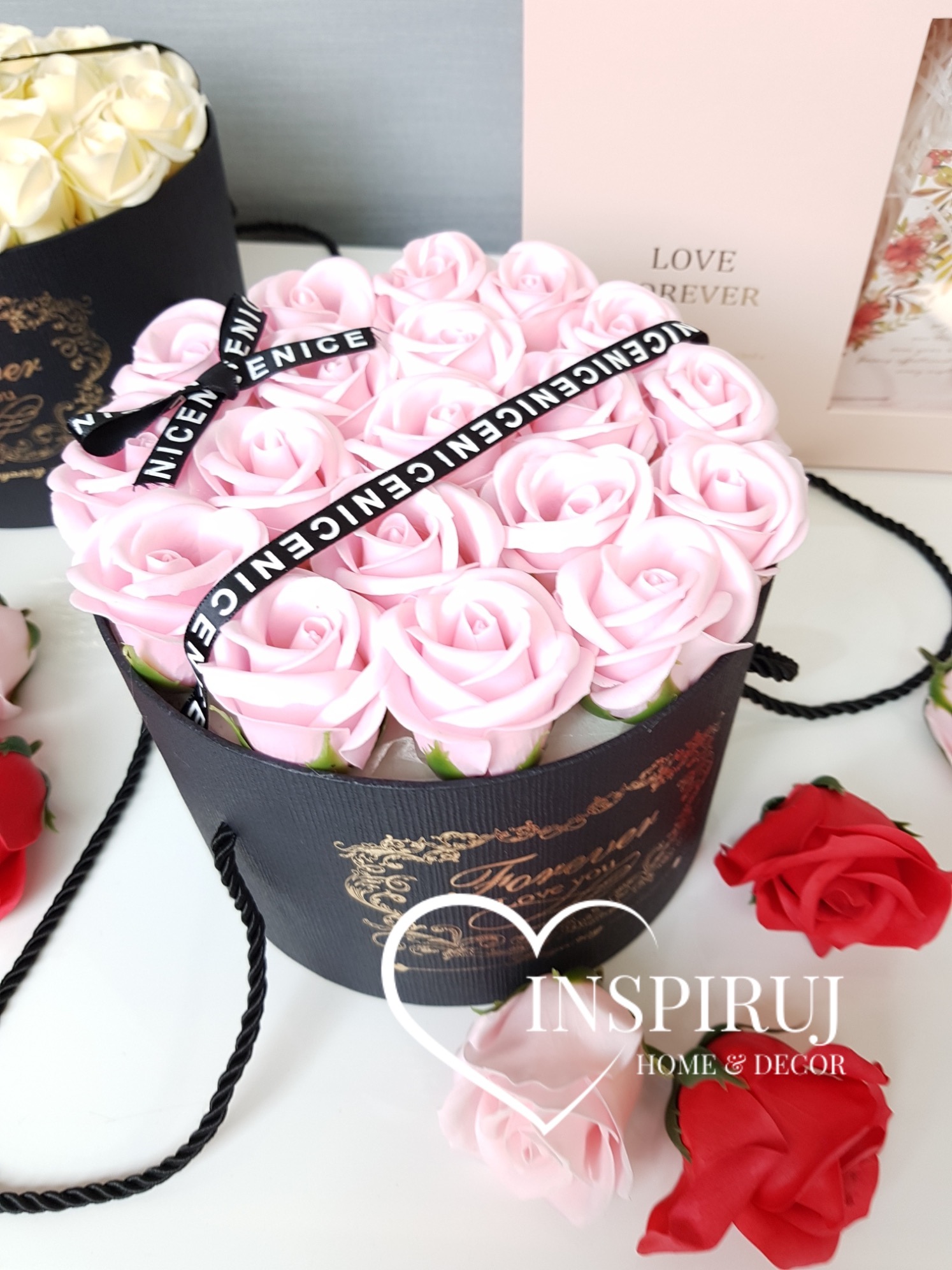 Różowe i czerwone mydlane róże w czarnych, ozdobnych pudełkach z uchwytami sznurkowymi, przewiązane wstążką z napisem.