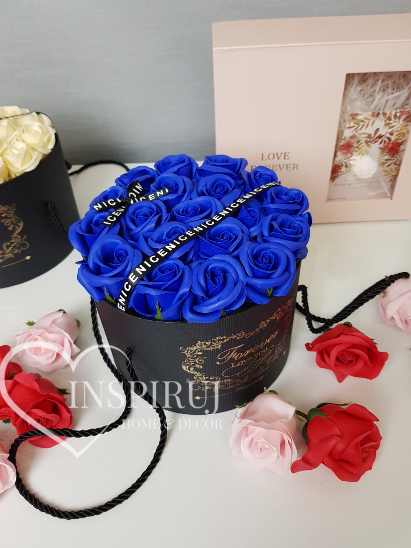 Kompozycja z niebieskich róż mydlanych w czarnym, okrągłym pudełku z napisem Forever, otoczona pojedynczymi różami w odcieniach czerwieni i różu. W tle widoczne inne pudełka.