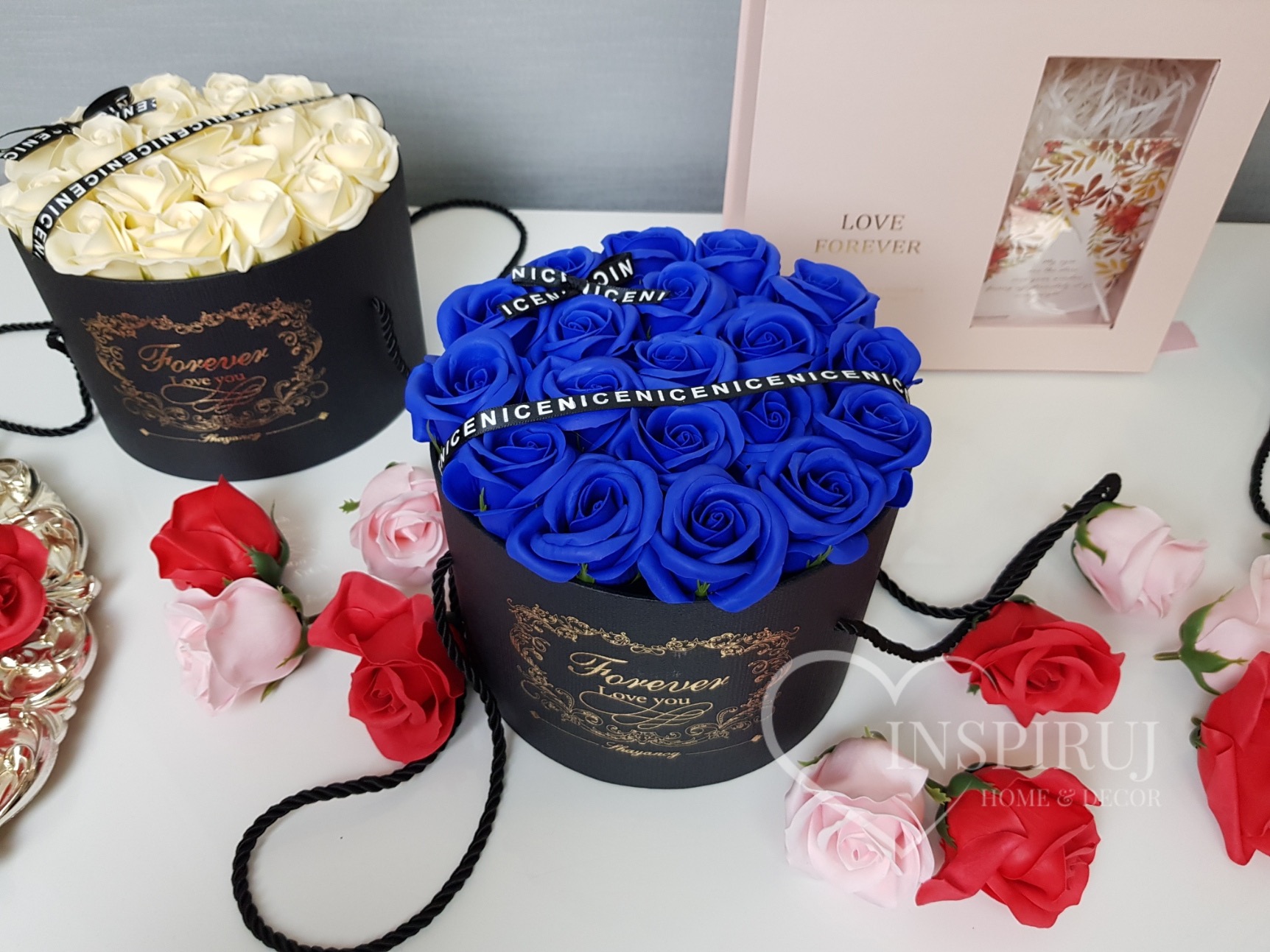 Dwa okrągłe pudełka z kwiatami w kolorach białym i niebieskim, otoczone pojedynczymi różami w odcieniach różu i czerwieni, prezentowane w Warszawie. Pudełka ozdobione napisem 'Forever Love You'...