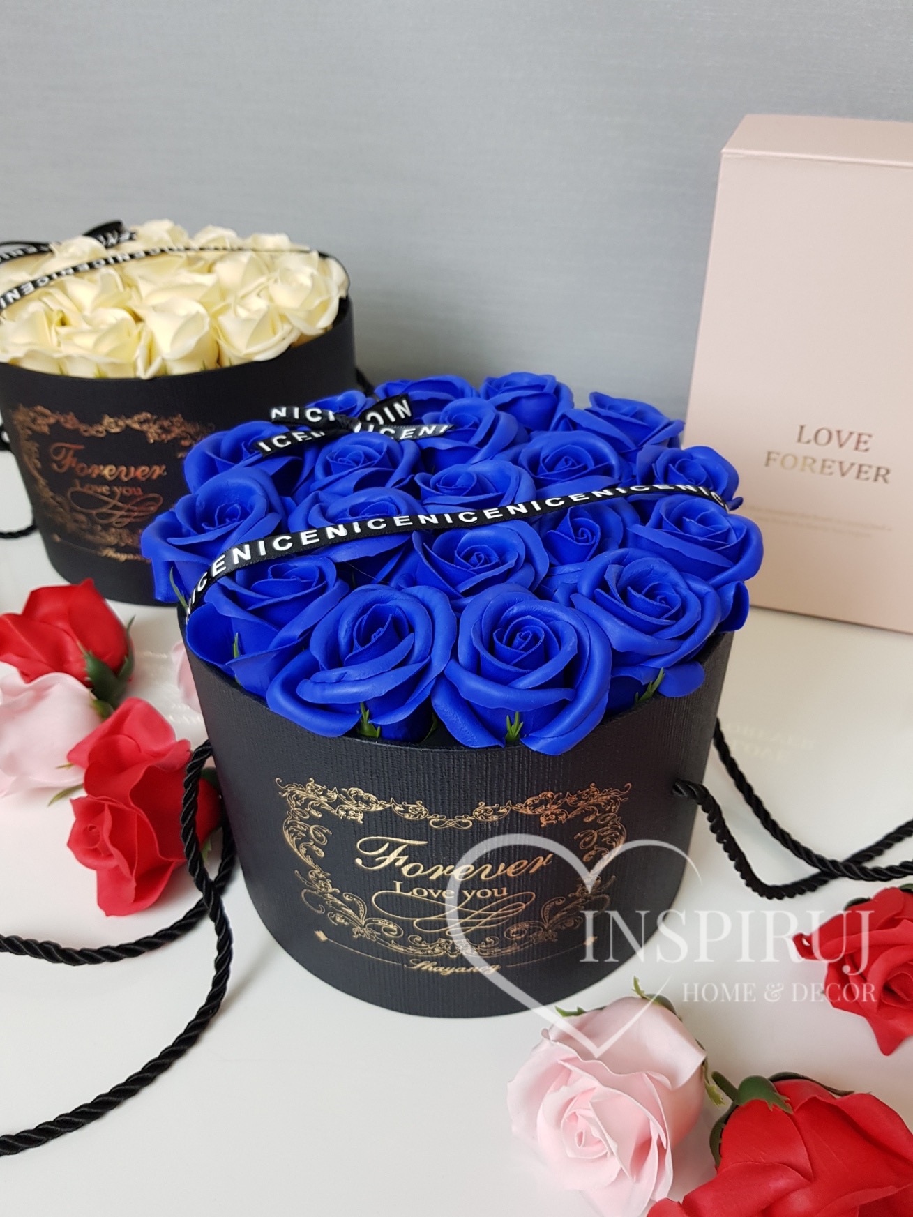 Kompozycja z niebieskich róż mydlanych w czarnym, okrągłym pudełku z napisem 'Forever Love you', otoczona czarną wstążką z powtarzającym się napisem, w tle pudełko z białymi różami i różowe pudełko...
