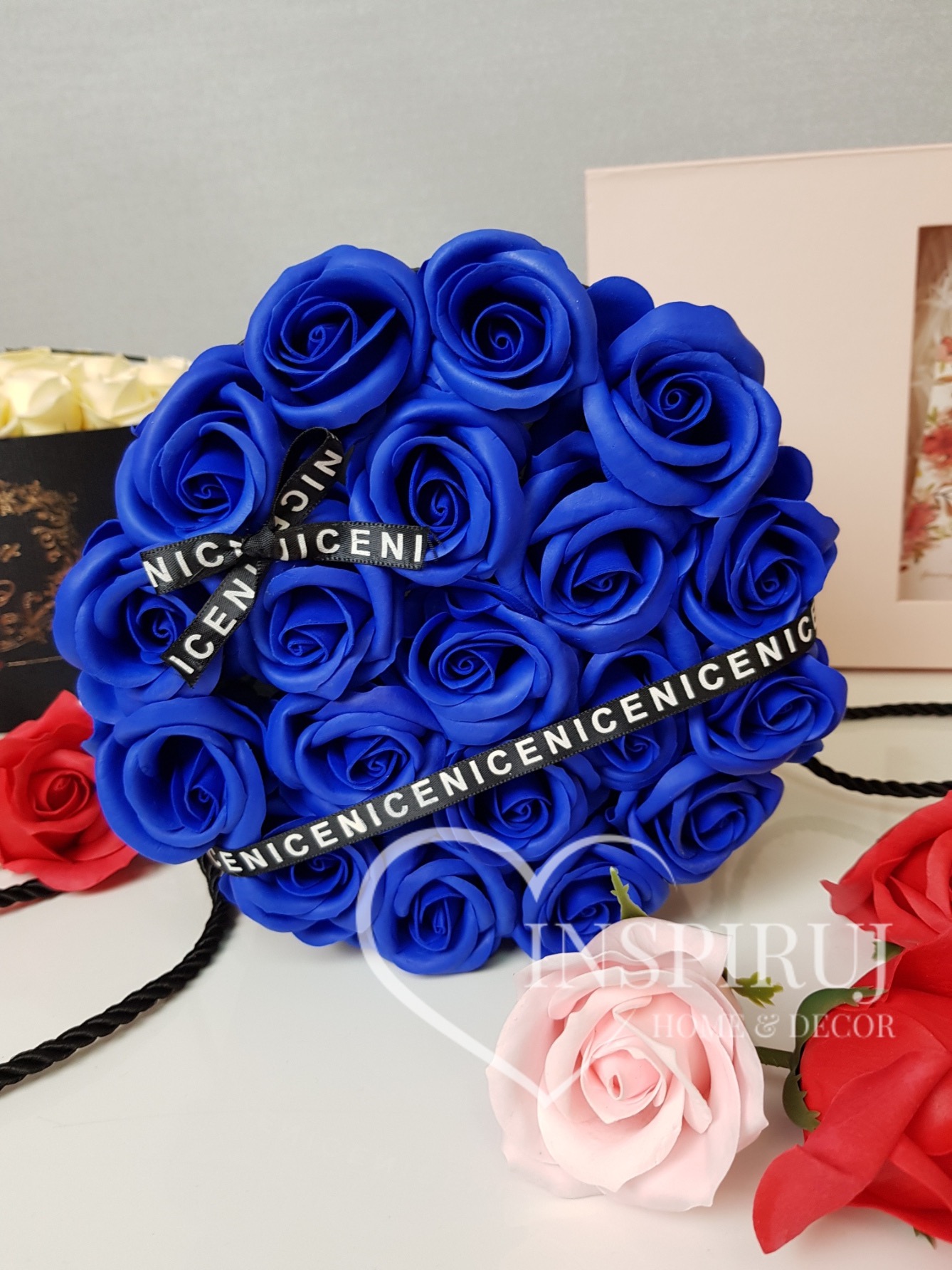 Bukiet z kobaltowych róż z czarną wstążką z napisem, otoczony pojedynczymi różami w odcieniach czerwieni i pudrowego różu, na jasnym tle.