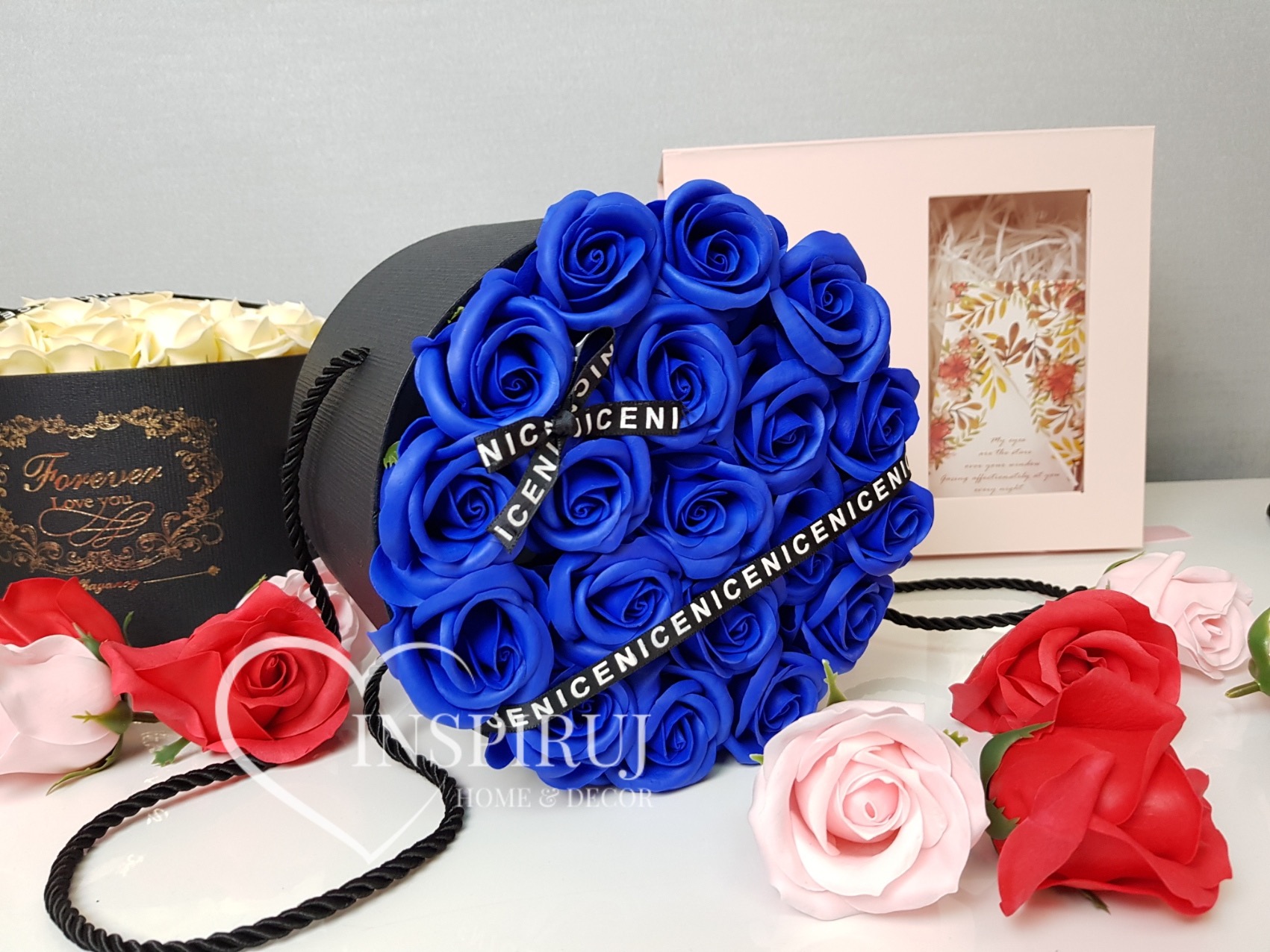 Kompozycja z niebieskich róż w czarnym pudełku z napisem 'NICENICENI', otoczona różowymi i czerwonymi różami, obok pudełko z białymi różami i ozdobne pudełko z życzeniami.