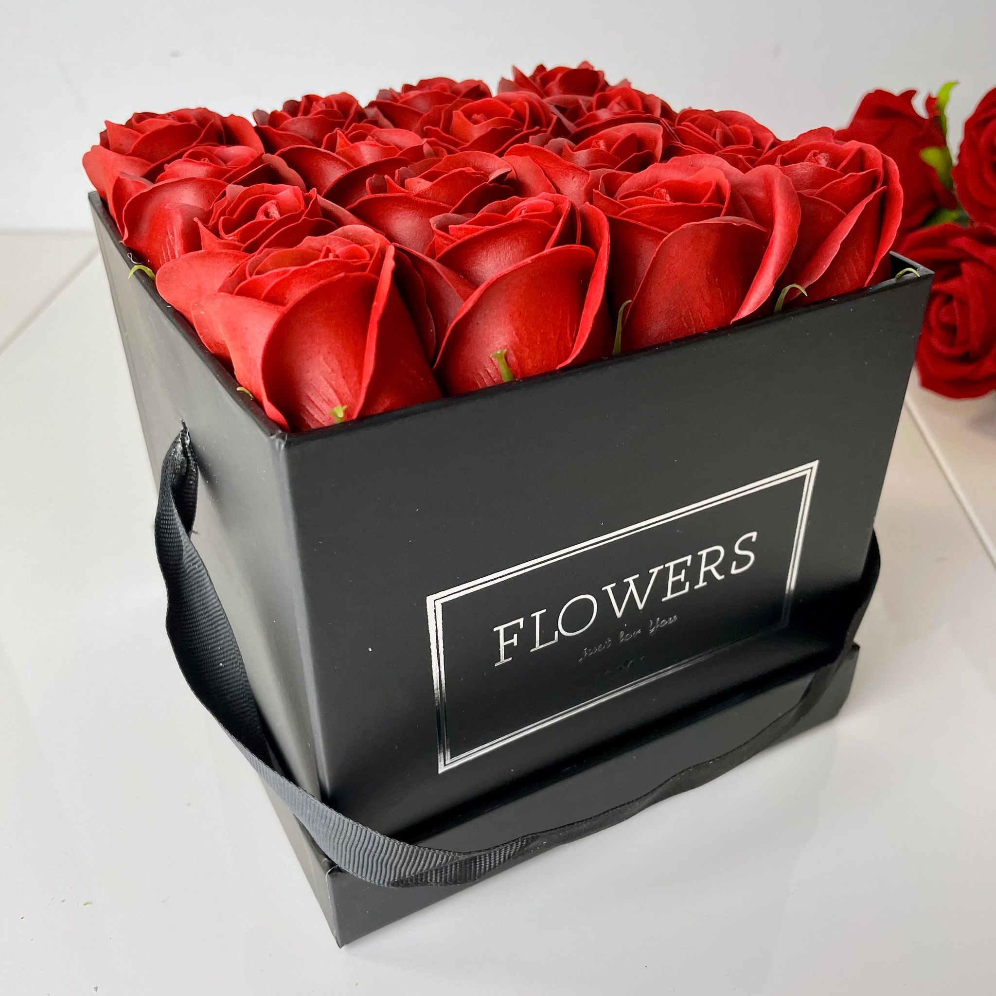 Kwadratowe, czarne pudełko z uchwytami wypełnione czerwonymi różami, widoczny napis 'FLOWERS' na boku.