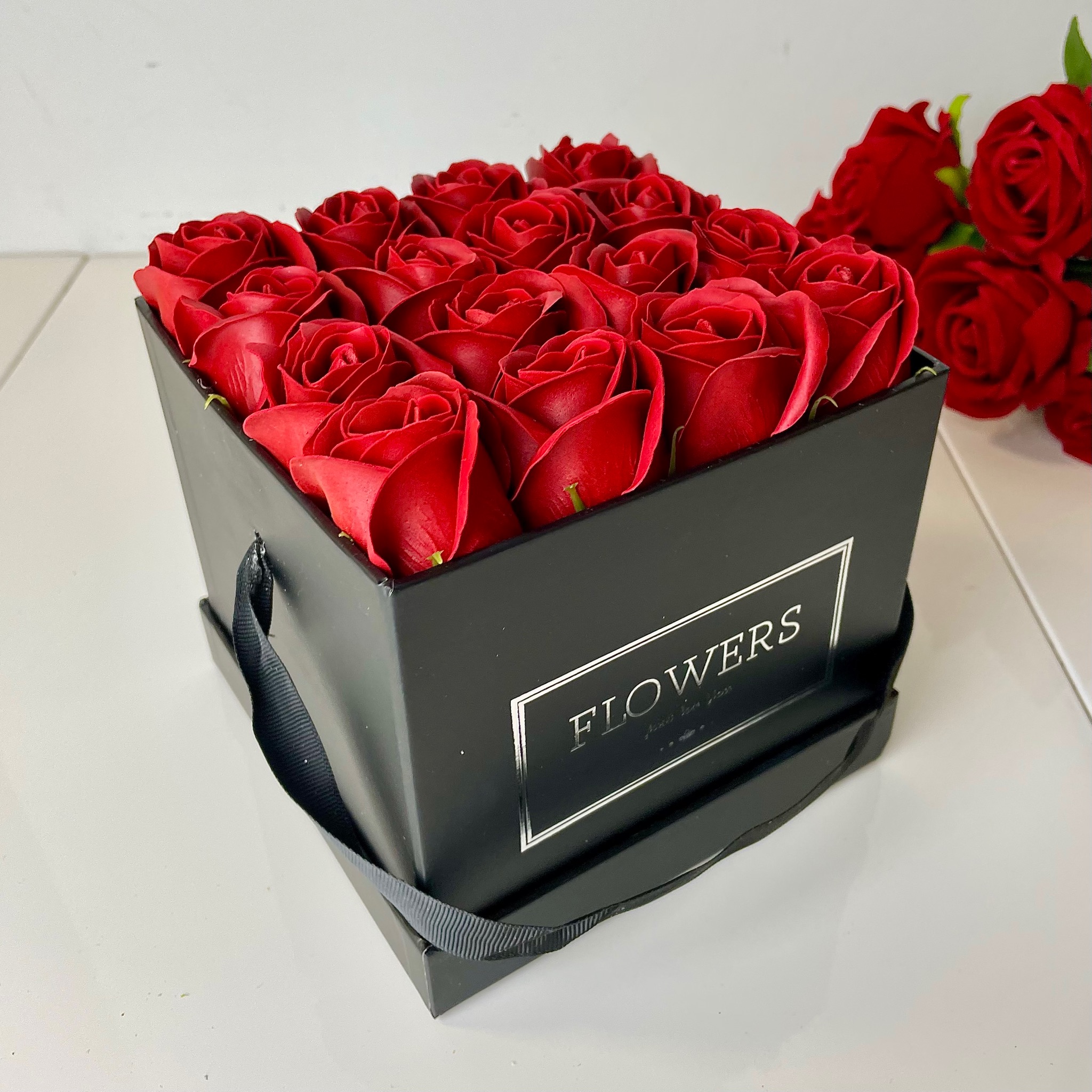 Kwadratowe, czarne pudełko z napisem 'FLOWERS' wypełnione czerwonymi różami, z dodatkowymi różami widocznymi w tle.