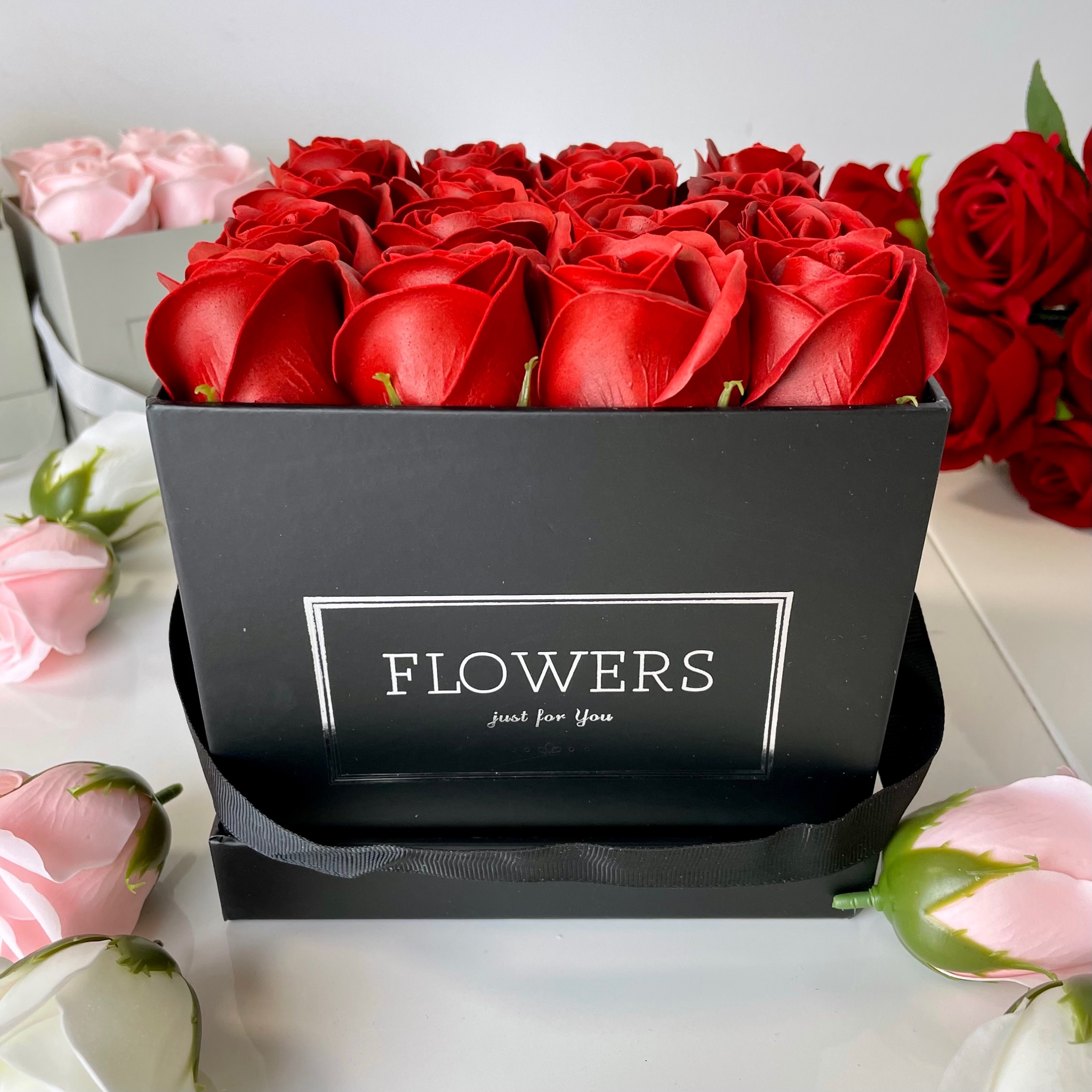 Kwadratowe, czarne pudełko wypełnione czerwonymi, sztucznymi różami, z napisem 'FLOWERS just for You', otoczone różowymi i białymi różami na białym tle.