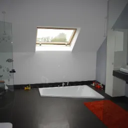 Nowoczesna łazienka na poddaszu z asymetryczną wanną, przeszklonym prysznicem z dekoracyjnymi kropkami, oknem dachowym i pomarańczowym dywanikiem, widziana z perspektywy wejścia.