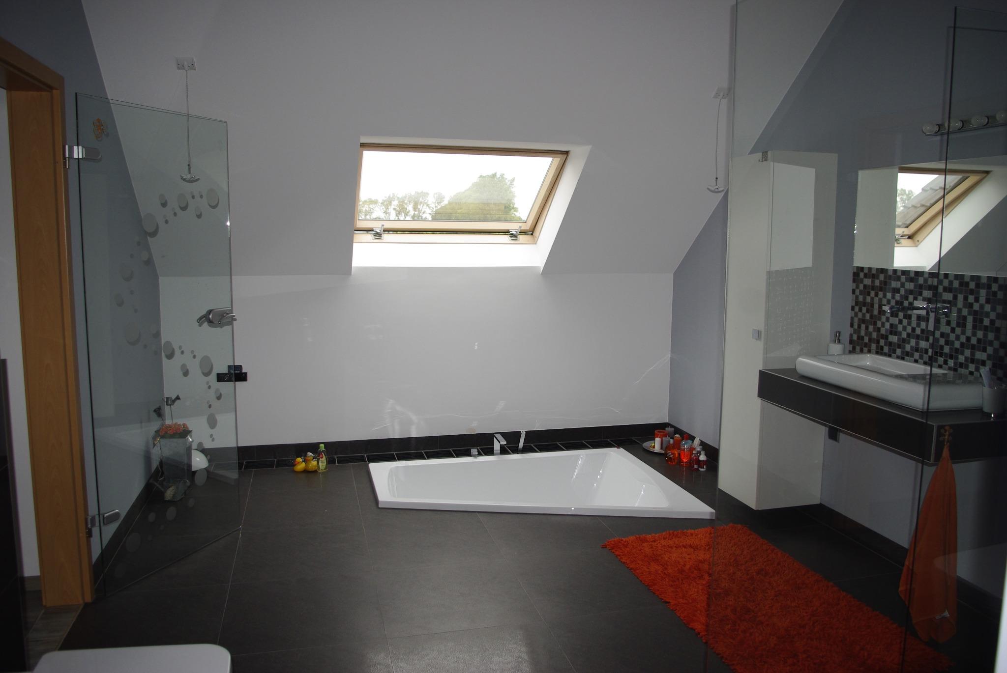 Nowoczesna łazienka na poddaszu z asymetryczną wanną, przeszklonym prysznicem z dekoracyjnymi kropkami, oknem dachowym i pomarańczowym dywanikiem, widziana z perspektywy wejścia.