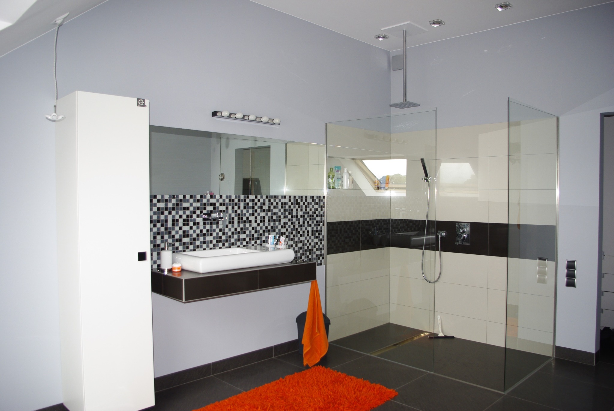 Nowoczesna łazienka z kabiną prysznicową typu walk-in, umywalką nablatową na ciemnym blacie, mozaiką za umywalką i pomarańczowym dywanikiem na ciemnej podłodze.