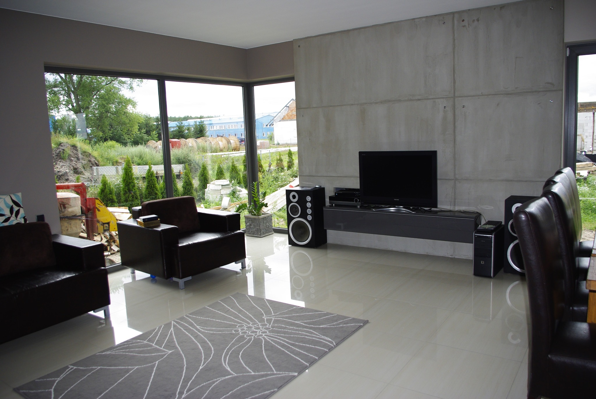 Nowoczesny salon z szarym dywanem w kwiaty, ciemnobrązowymi skórzanymi fotelami i betonową ścianą za telewizorem, widok na ogród przez duże okno.