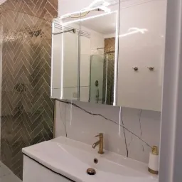 Nowoczesna łazienka z białymi meblami, złotą armaturą i lustrem z podświetleniem LED, widoczna kabina prysznicowa z mozaiką w jodełkę.