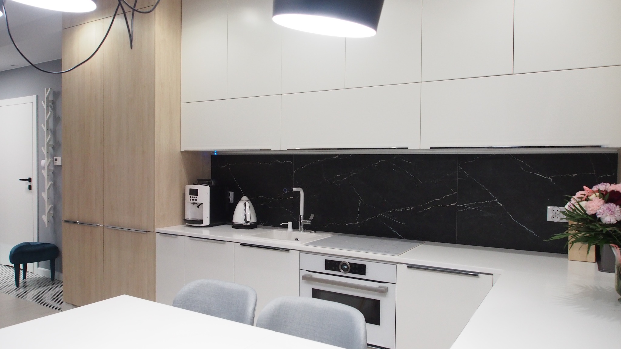 Nowoczesna kuchnia z białymi szafkami, czarnym marmurowym backsplashiem, białymi sprzętami i drewnianą szafą po lewej stronie, uzupełniona bukietem kwiatów po prawej stronie.