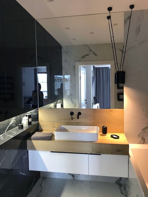 Nowoczesna łazienka z marmurową ścianą i blatem, białą umywalką nablatową oraz czarną armaturą i lampami wiszącymi na tle dużego lustra.