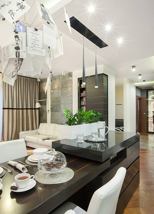 Nowoczesny salon z aneksem kuchennym: stół z nakryciem, szklany klosz z ciastkami, minimalistyczne lampy wiszące, ściana z szarej cegły i sofa w tle.