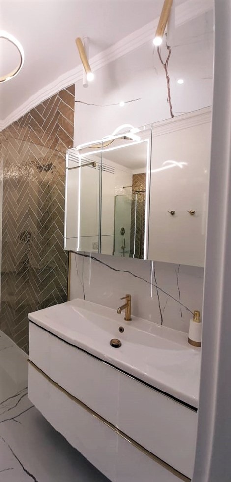 Nowoczesna łazienka z białymi meblami, złotą armaturą i lustrem z podświetleniem LED, widoczna kabina prysznicowa z mozaiką w jodełkę.