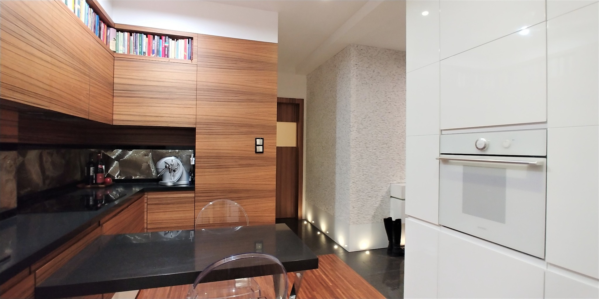 Nowoczesna kuchnia z drewnianymi szafkami, książkami na górnej półce, czarnym blatem, przezroczystymi krzesłami i białym piekarnikiem w zabudowie.