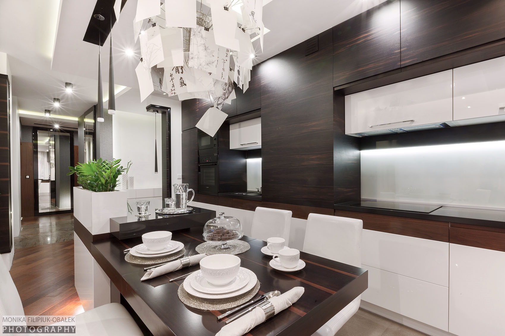 Nowoczesny salon z aneksem kuchennym w ciemnych barwach drewna i bieli, stół z nakryciem, designerski żyrandol z papierowymi elementami, widok na korytarz z roślinami.