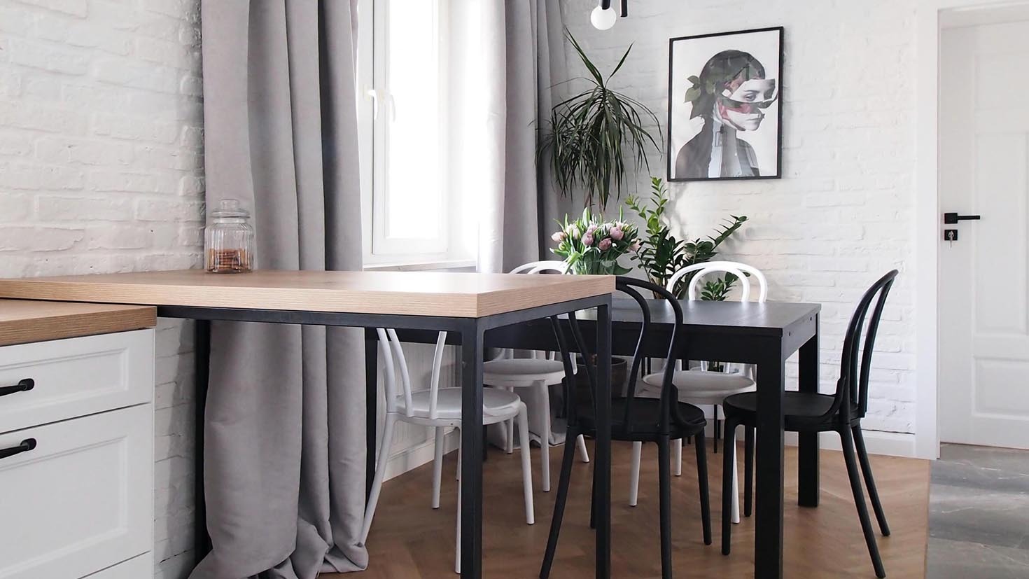 Jasna kuchnia z drewnianym blatem, czarnym stołem i krzesłami w stylu skandynawskim, z białymi ścianami z cegły i minimalistycznym plakatem.
