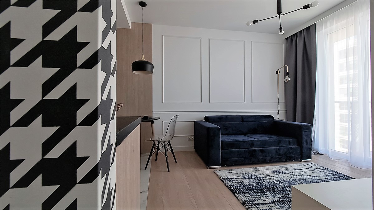 Wnętrze kawalerki z ciemnoniebieską sofą, wzorzystą ścianą w pepitkę i przezroczystym krzesłem, oświetlone lampami wiszącymi i stojącą