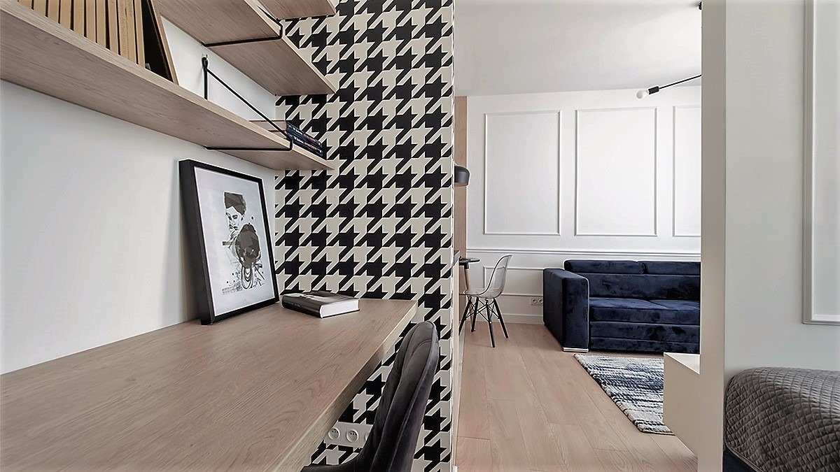 Nowoczesna kawalerka z biurkiem, półkami, tapetą w pepitkę i granatową sofą w tle.