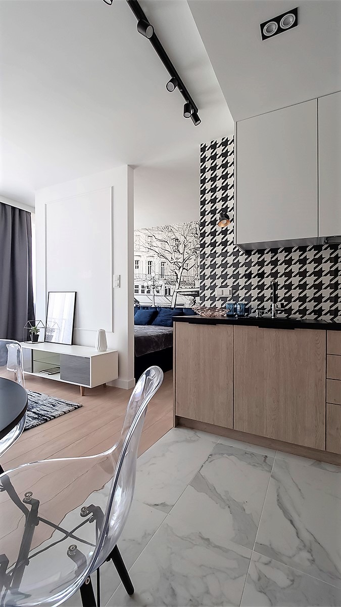 Wnętrze kawalerki z aneksem kuchennym: białe ściany, podłoga z jasnego drewna, marmurowe płytki, meble w odcieniach bieli i drewna, ściana z wzorem w pepitkę, przezroczyste krzesło, w tle widoczny...