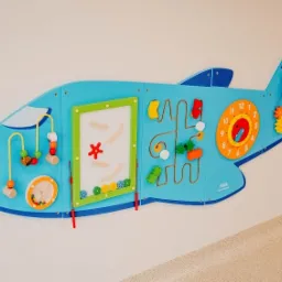 Niebieska tablica manipulacyjna w kształcie samolotu, zamontowana na ścianie, z elementami edukacyjnymi i sensorycznymi dla dzieci, takimi jak labirynt, zegar i koraliki.