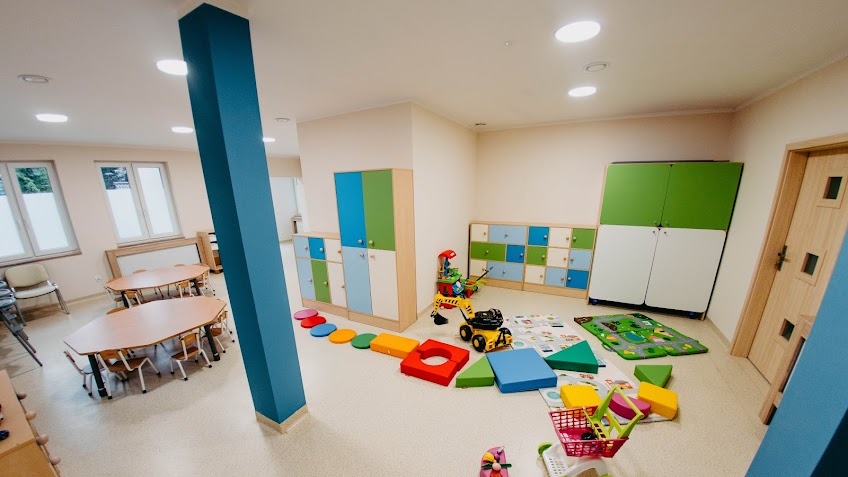 Jasne wnętrze sali zabaw z kolorowymi szafkami, stolikami i krzesłami dla dzieci, oraz różnorodnymi zabawkami edukacyjnymi i interaktywnymi.