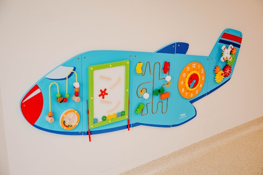Niebieska tablica manipulacyjna w kształcie samolotu, zamontowana na ścianie, z elementami edukacyjnymi i sensorycznymi dla dzieci, takimi jak labirynt, zegar i koraliki.