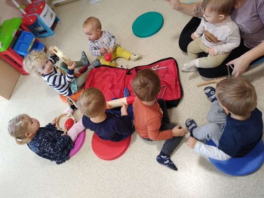 Grupa dzieci siedzi na kolorowych poduszkach, bawiąc się instrumentami muzycznymi, w tle widoczna torba na instrumenty i zabawki.