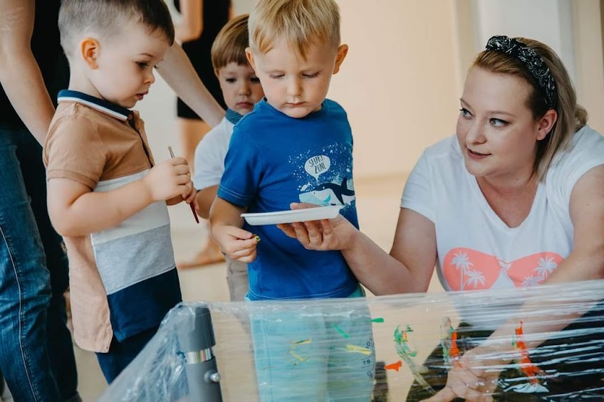 Grupa dzieci maluje farbami na folii pod nadzorem opiekunki, widoczne pędzle i paleta z farbami.