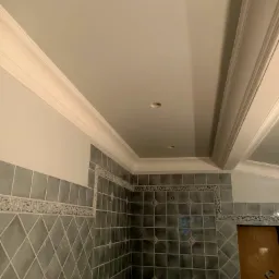 Świeżo wykafelkowana ściana łazienki z dekoracyjnym pasem płytek i listwą przysufitową, widoczne otwory na oświetlenie w suficie, częściowo zakryte wejście do wnęki.