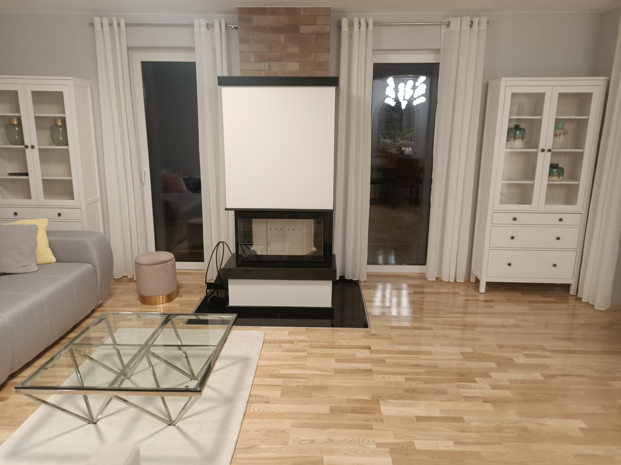 Jasny salon z parkietem, kominkiem obudowanym cegłą i białymi meblami, widok na ogród przez okno.