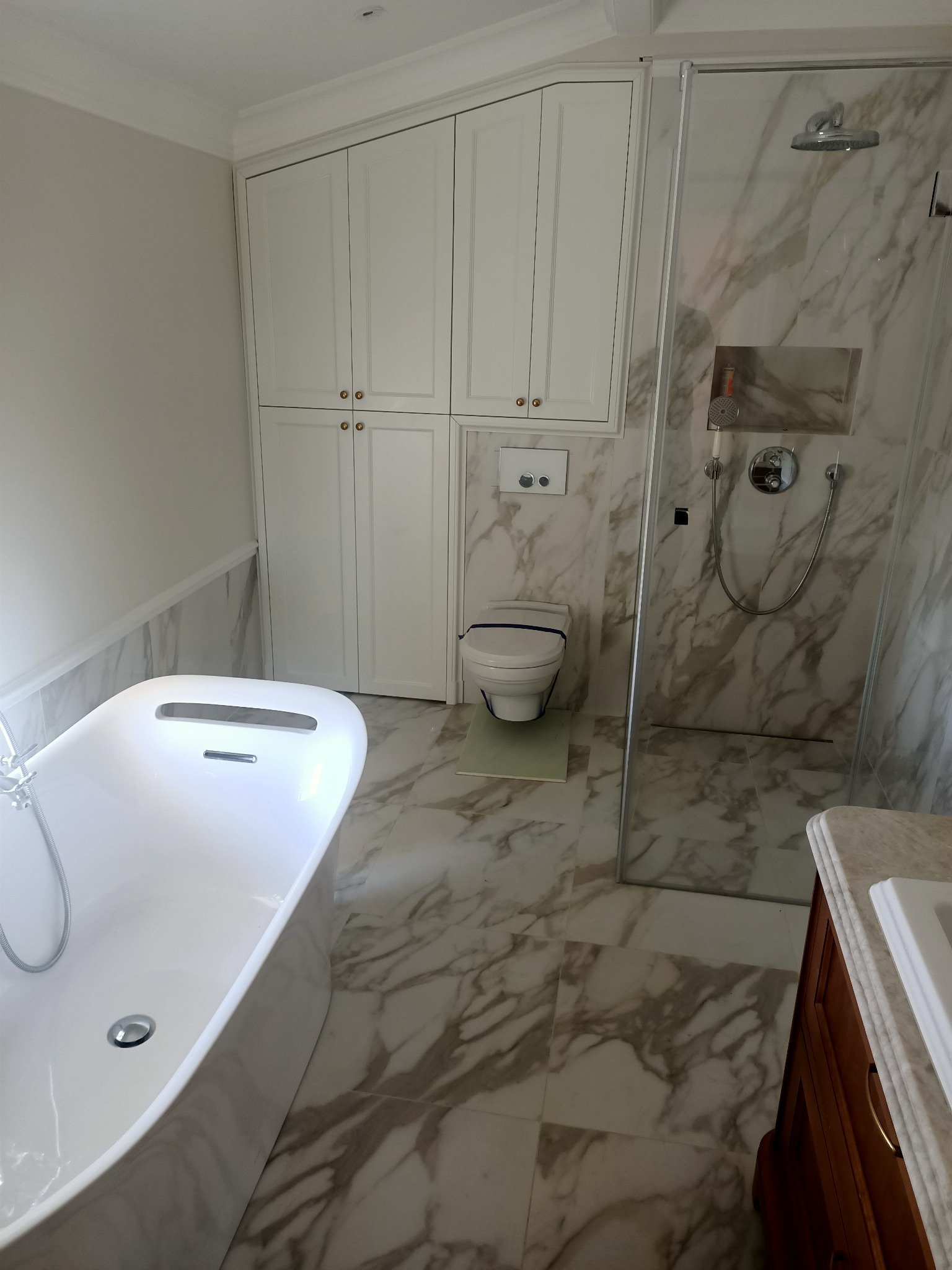 Nowoczesna łazienka z marmurowymi płytkami, kabiną prysznicową ze szklanymi drzwiami, wolnostojącą wanną i białymi szafkami wiszącymi.