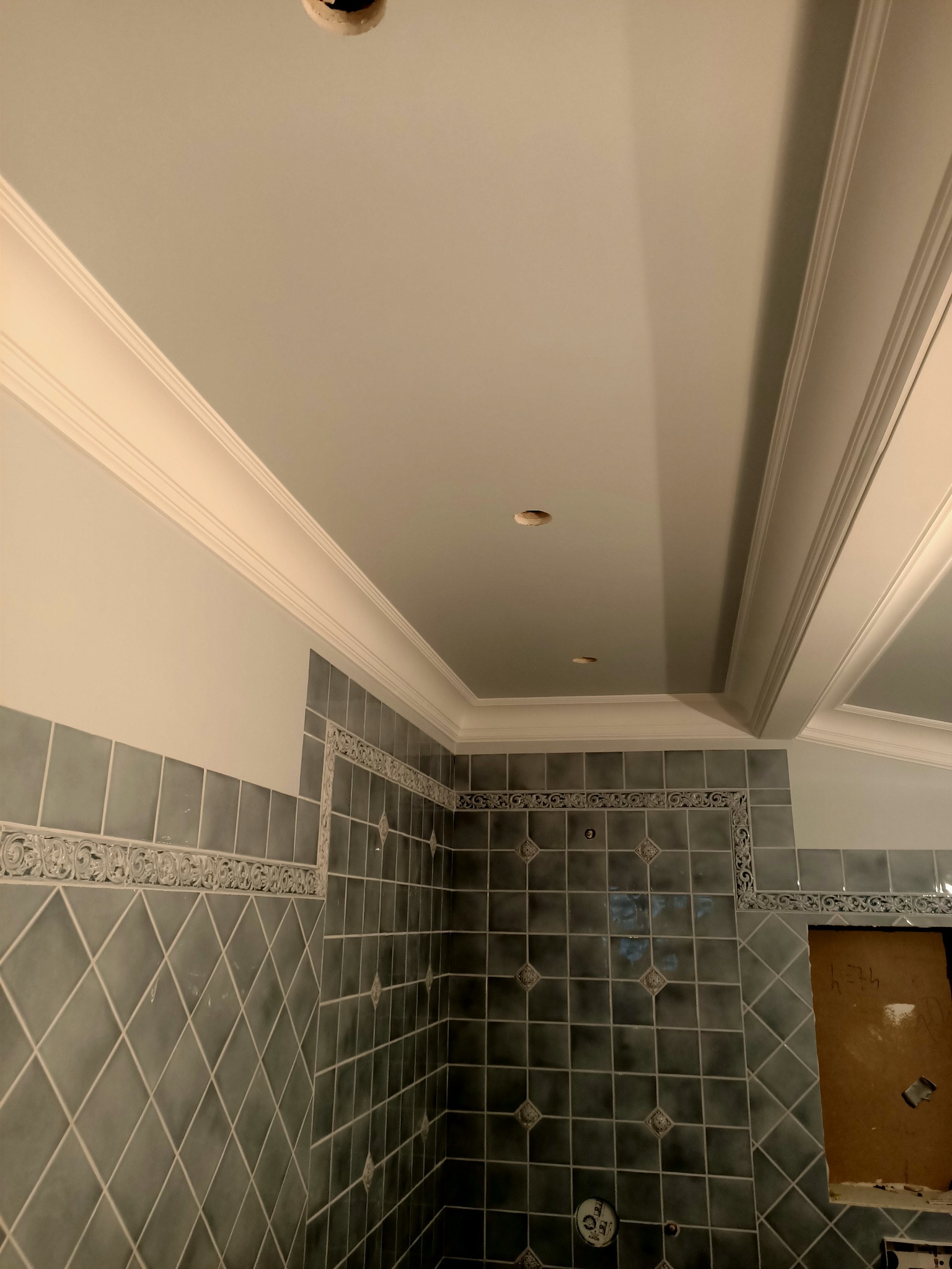 Świeżo wykafelkowana ściana łazienki z dekoracyjnym pasem płytek i listwą przysufitową, widoczne otwory na oświetlenie w suficie, częściowo zakryte wejście do wnęki.