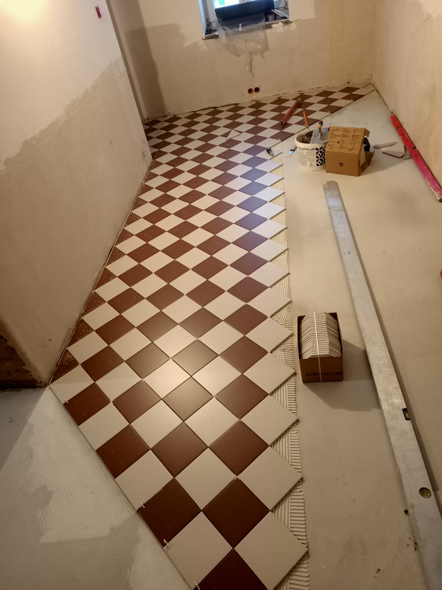 Układanie brązowo-białej szachownicy z płytek podłogowych w trakcie prac, widoczne narzędzia glazurnicze, poziomica i paczka z płytkami.