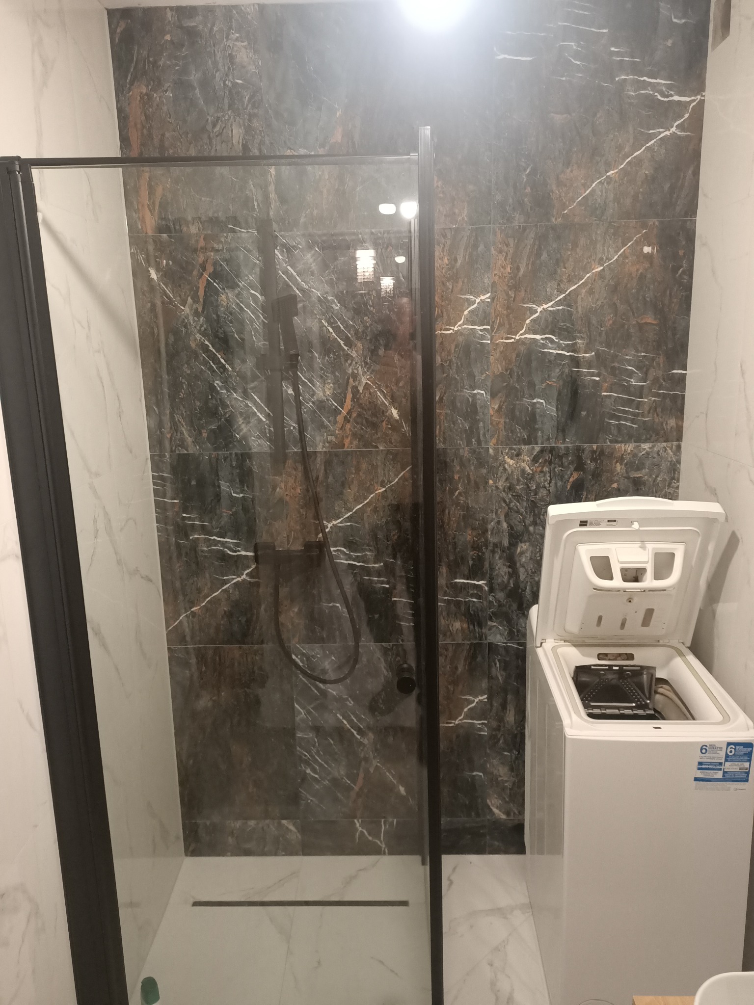 Nowoczesna łazienka z kabiną prysznicową z czarnymi ramami i marmurowymi płytkami w odcieniach szarości i brązu, obok stoi biała pralka z otwartą klapą.