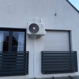 Thermopol - Zewnętrzna jednostka klimatyzatora Rotenso Unico zamontowana na jasnoszarej elewacji budynku, między oknem z zasłonami a oknem z roletą, z ciemnymi balustradami poniżej.