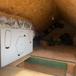 Thermopol - Biały rekuperator THESLAGREEN z podłączonym srebrnym kanałem wentylacyjnym, umieszczony na podłodze z płyt OSB w niezagospodarowanym poddaszu. W tle widoczne inne kanały i prowizoryczne oświetlenie.