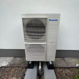 Zewnętrzna jednostka klimatyzacji Panasonic zamontowana na betonowych podstawach przy ścianie budynku z widoczną izolacją rur.