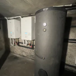 Dwie białe jednostki grzewcze Panasonic zamontowane na szarej ścianie, obok pionowego, cylindrycznego zbiornika Aquatec Systems z widocznym manometrem.