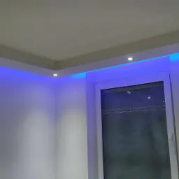 Białe wnętrze z podwieszanym sufitem, oświetleniem punktowym i dekoracyjnym niebieskim światłem LED wokół sufitu i okna.