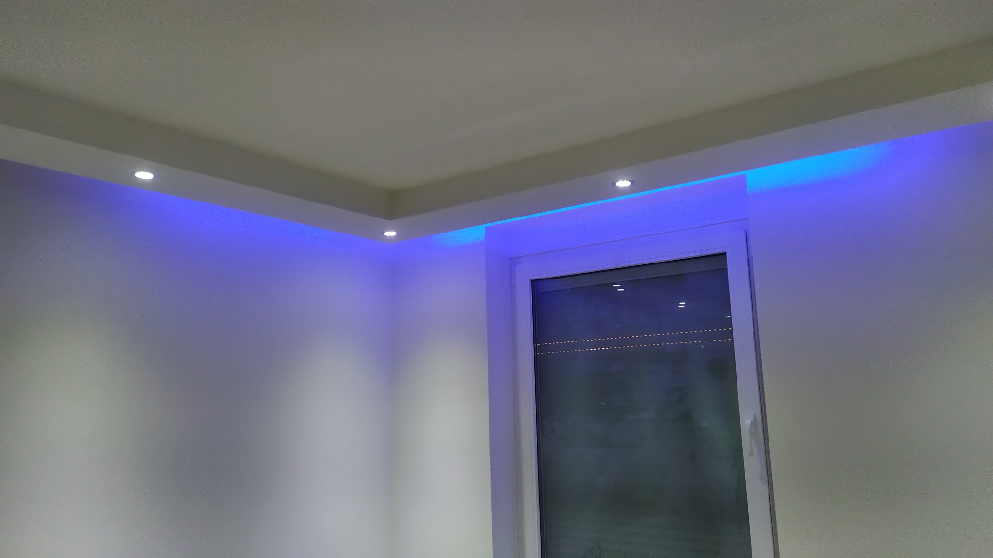 Białe wnętrze z podwieszanym sufitem, oświetleniem punktowym i dekoracyjnym niebieskim światłem LED wokół sufitu i okna.