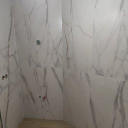 BUD CORP - Świeżo wykafelkowana ściana łazienki z białymi płytkami imitującymi marmur, widoczne otwory na armaturę, drewnopodobna podłoga, surowe wykończenie sufitu.