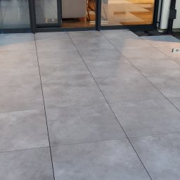 BUD CORP - Jasnoszare płytki ułożone na zewnątrz, przy drzwiach tarasowych, poziomica leżąca obok, widoczny fragment izolacji.